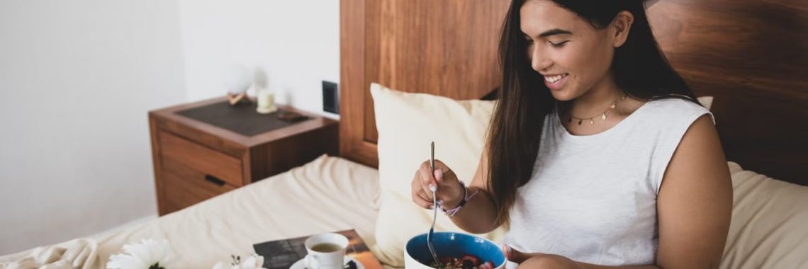 Woman enjoying breakfast in bed with a bowl of granola and fruit, creating a cozy atmosphere.